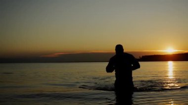 Gömlekli vahşi bir adamın silueti gün batımında denizden sahile çıkar. Adam sudan çıkıyor..