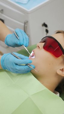 Dişçi muayenehanesindeki kız çocuğu. Modern bir diş kliniğinde küçük bir kızın dişlerini tedavi eden dişçilerin yakın çekimi. Çocukların dişlerinin tedavisi için. Dikey video.