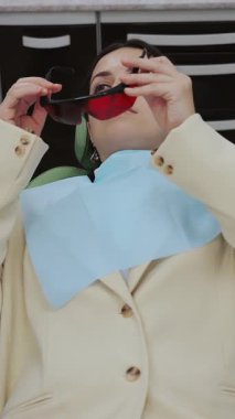 Diş hekimi koltuğundaki bir kadın hasta diş tedavisinden önce koruyucu gözlük takar. Modern diş kliniği. Diş tedavisi. Dikey video.