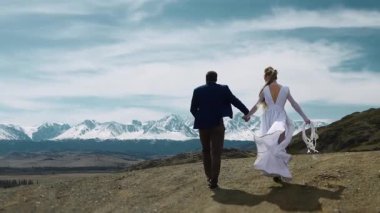 A newlywed man and woman in wedding dress walking along a mountain top against the backdrop of snow-capped mountain peaks.