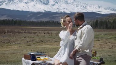 A young couple on a picnic in the mountains, against the backdrop of snow-capped peaks. A man and woman kiss while traveling.