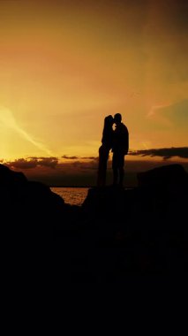 A silhouette of two people in love, a man and a woman, standing on the seashore at sunset. Family vacation on a tropical coast. Vertical video.