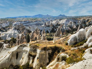 Manzara, Cappadocia 'nın ikonik kaya oluşumlarını ve kayan tepeleri, yemyeşil ve parlak gökyüzünün altında görünen uzak tarihi yerleri sergiliyor..