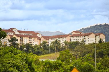 Kırmızı kiremitli çatıları olan bir apartman dairesi yığını. Bereketli yeşillik, ağaçlar ve tepeler bulutlu mavi gökyüzünün altında onları çevreleyen Kuala Lumpur banliyö bölgesinde.