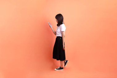 Tayland üniversitesi üniformalı neşeli bir bayan öğrenci. Portakal rengi arka planda cep telefonu ve kulaklık taşıyormuş. Uluslararası Öğrenciler Günü, Dünya Öğrenciler Günü. Gen Z