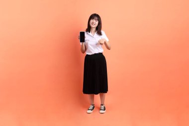 Tayland Üniversitesi üniformalı neşeli bayan öğrenci portakal rengi arka planda izole edilmiş cep telefonunu tutuyor. Uluslararası Öğrenciler Günü, Dünya Öğrenciler Günü. Gen Z