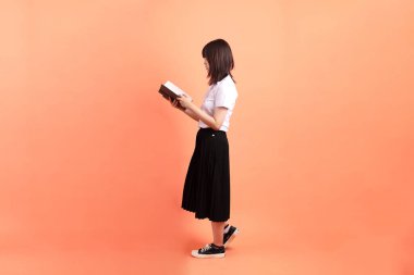 Tayland üniversitesi üniformalı neşeli bir bayan öğrenci. Elinde turuncu arka planda izole edilmiş bir ders kitabı var. Uluslararası Öğrenciler Günü, Dünya Öğrenciler Günü. Gen Z