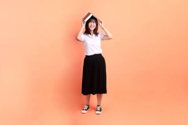 Tayland üniversitesi üniformalı neşeli bir bayan öğrenci. Elinde turuncu arka planda izole edilmiş bir ders kitabı var. Uluslararası Öğrenciler Günü, Dünya Öğrenciler Günü. Gen Z