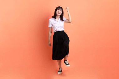 Tayland üniversitesi üniformalı neşeli bayan öğrenci turuncu arka planda izole edilmiş yürüyüş jestleriyle. Uluslararası Öğrenciler Günü, Dünya Öğrenciler Günü. Gen Z