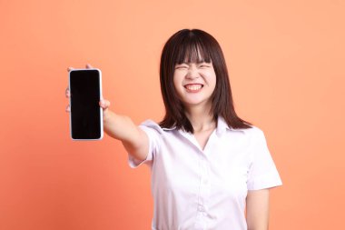 Tayland Üniversitesi üniformalı neşeli bayan öğrenci portakal rengi arka planda izole edilmiş cep telefonunu tutuyor. Uluslararası Öğrenciler Günü, Dünya Öğrenciler Günü. Gen Z