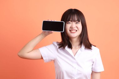 Tayland Üniversitesi üniformalı neşeli bayan öğrenci portakal rengi arka planda izole edilmiş cep telefonunu tutuyor. Uluslararası Öğrenciler Günü, Dünya Öğrenciler Günü. Gen Z