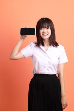 Tayland Üniversitesi üniformalı neşeli bayan öğrenci portakal rengi arka planda izole edilmiş cep telefonunu tutuyor. Uluslararası Öğrenciler Günü, Dünya Öğrenciler Günü. Gen Z