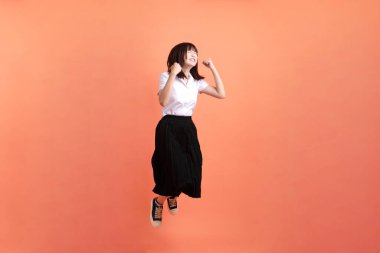 Tayland üniversitesi üniformalı neşeli bayan öğrenci turuncu arka planda izole atlamanın hareketleriyle. Uluslararası Öğrenciler Günü, Dünya Öğrenciler Günü. Gen Z