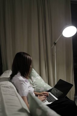 Tayland üniversitesi üniformalı genç bir kız öğrenci oturma odasındaki bir kanepede oturur ve bir dizüstü bilgisayarla internetten ders çalışır. Uluslararası Öğrenciler Günü, Dünya Öğrenciler Günü. Gen Z, E-öğrenme