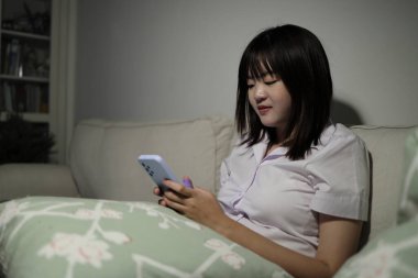 Tayland üniversitesi üniformalı genç bir kız öğrenci oturma odasında bir kanepede oturuyor, cep telefonuyla internetten çalışıyor. Uluslararası Öğrenciler Günü, Gen Z, E-öğrenim