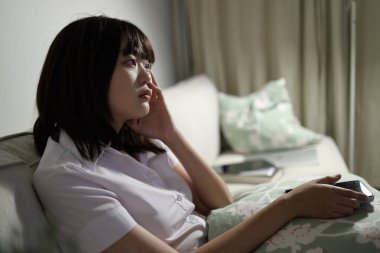 Tayland üniversitesi üniformalı genç bir kız öğrenci oturma odasında bir kanepede oturuyor, cep telefonuyla internetten çalışıyor. Uluslararası Öğrenciler Günü, Gen Z, E-öğrenim