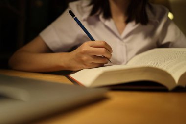 Tayland üniversitesi üniformalı genç bir kız öğrenci çalışma masasında oturur, ödev yapar ve çok çalışır. Uluslararası Öğrenciler Günü, Dünya Öğrenciler Günü. Gen Z, E-öğrenme