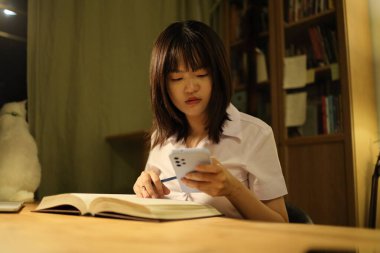 Tayland üniversitesi üniformalı genç bir kız öğrenci çalışma masasında oturur, ödev yapar ve çok çalışır. Uluslararası Öğrenciler Günü, Dünya Öğrenciler Günü. Gen Z, E-öğrenme