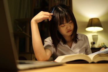 Tayland üniversitesi üniformalı genç bir kız öğrenci çalışma masasında oturur, ödev yapar ve çok çalışır. Uluslararası Öğrenciler Günü, Dünya Öğrenciler Günü. Gen Z, E-öğrenme