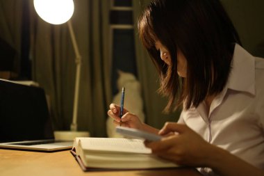 Tayland üniversitesi üniformalı genç bir kız öğrenci çalışma masasında oturur, ödev yapar ve çok çalışır. Uluslararası Öğrenciler Günü, Dünya Öğrenciler Günü. Gen Z, E-öğrenme