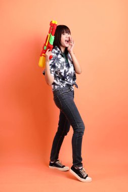 Mutlu SONGKRAN GÜNÜ. Turuncu arka planda plastik su tabancası tutan yaz kıyafetleri içinde neşeli, sevimli, genç Asyalı bir kadın. Songkran festivali. Tayland Yeni Yıl Günü.