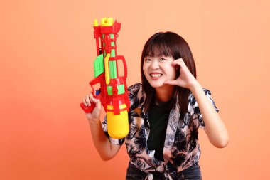 Mutlu SONGKRAN GÜNÜ. Turuncu arka planda plastik su tabancası tutan yaz kıyafetleri içinde neşeli, sevimli, genç Asyalı bir kadın. Songkran festivali. Tayland Yeni Yıl Günü.