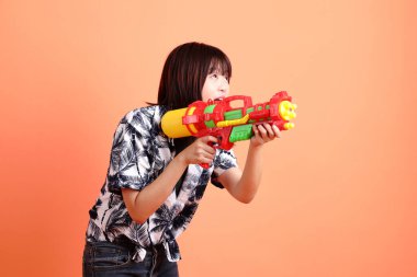 Mutlu SONGKRAN GÜNÜ. Turuncu arka planda plastik su tabancası tutan yaz kıyafetleri içinde neşeli, sevimli, genç Asyalı bir kadın. Songkran festivali. Tayland Yeni Yıl Günü.