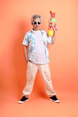 Mutlu SONGKRAN GÜNÜ. Yazlık kıyafetleri ve güneş gözlükleriyle turuncu arka planda izole edilmiş plastik su tabancası tutan Asyalı enerjik kıdemli bir adam. Songkran festivali. Tayland Yeni Yıl Günü.