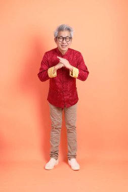 Yeni Çin yılın kutlu olsun. Asya 'nın enerjik kıdemli adamı kırmızı geleneksel kıyafetler giyiyor ve turuncu arka planda izole edilmiş bir selamlama hareketi yapıyor..