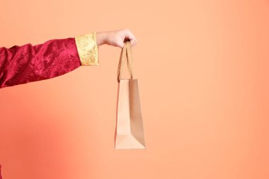 Yeni Çin yılın kutlu olsun. Turuncu arka planda izole edilmiş kahverengi kağıt poşeti tutan kırmızı geleneksel kıyafetler giyen Asyalı son sınıf öğrencisinin eli..