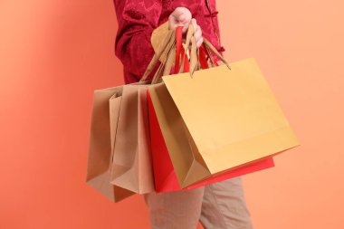 Yeni Çin yılın kutlu olsun. Turuncu arka planda izole edilmiş kağıt poşetlerle el ele tutuşan geleneksel kırmızı elbiseler giyen Asyalı son sınıf öğrencisinin eli..