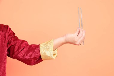 Yeni Çin yılın kutlu olsun. Kırmızı geleneksel cheongsam qipao ya da changshan elbisesi giyen Asyalı son sınıf öğrencisinin eli turuncu arka planda izole edilmiş gümüş çubuklarla el ele tutuşuyor..