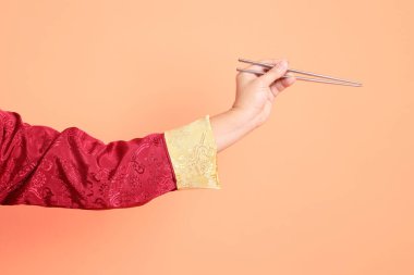 Yeni Çin yılın kutlu olsun. Kırmızı geleneksel cheongsam qipao ya da changshan elbisesi giyen Asyalı son sınıf öğrencisinin eli turuncu arka planda izole edilmiş gümüş çubuklarla el ele tutuşuyor..