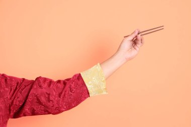 Yeni Çin yılın kutlu olsun. Kırmızı geleneksel cheongsam qipao ya da changshan elbisesi giyen Asyalı son sınıf öğrencisinin eli turuncu arka planda izole edilmiş gümüş çubuklarla el ele tutuşuyor..