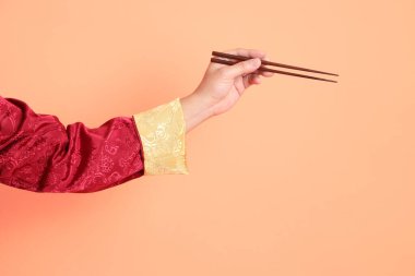 Yeni Çin yılın kutlu olsun. Kırmızı geleneksel cheongsam qipao ya da changshan elbisesi giyen Asyalı son sınıf öğrencisinin eli turuncu arka planda izole edilmiş kahverengi çubukları tutan el hareketleriyle..