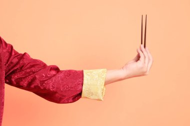 Yeni Çin yılın kutlu olsun. Kırmızı geleneksel cheongsam qipao ya da changshan elbisesi giyen Asyalı son sınıf öğrencisinin eli turuncu arka planda izole edilmiş kahverengi çubukları tutan el hareketleriyle..