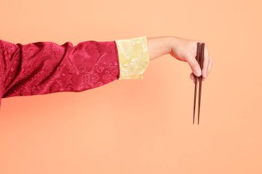 Yeni Çin yılın kutlu olsun. Kırmızı geleneksel cheongsam qipao ya da changshan elbisesi giyen Asyalı son sınıf öğrencisinin eli turuncu arka planda izole edilmiş kahverengi çubukları tutan el hareketleriyle..