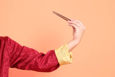 Yeni Çin yılın kutlu olsun. Kırmızı geleneksel cheongsam qipao ya da changshan elbisesi giyen Asyalı son sınıf öğrencisinin eli turuncu arka planda izole edilmiş kahverengi çubukları tutan el hareketleriyle..