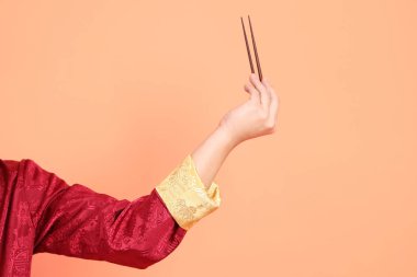 Yeni Çin yılın kutlu olsun. Kırmızı geleneksel cheongsam qipao ya da changshan elbisesi giyen Asyalı son sınıf öğrencisinin eli turuncu arka planda izole edilmiş kahverengi çubukları tutan el hareketleriyle..