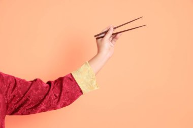 Yeni Çin yılın kutlu olsun. Kırmızı geleneksel cheongsam qipao ya da changshan elbisesi giyen Asyalı son sınıf öğrencisinin eli turuncu arka planda izole edilmiş kahverengi çubukları tutan el hareketleriyle..