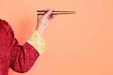 Yeni Çin yılın kutlu olsun. Kırmızı geleneksel cheongsam qipao ya da changshan elbisesi giyen Asyalı son sınıf öğrencisinin eli turuncu arka planda izole edilmiş kahverengi çubukları tutan el hareketleriyle..