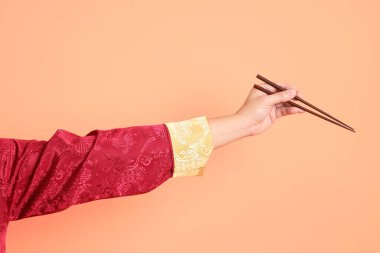 Yeni Çin yılın kutlu olsun. Kırmızı geleneksel cheongsam qipao ya da changshan elbisesi giyen Asyalı son sınıf öğrencisinin eli turuncu arka planda izole edilmiş kahverengi çubukları tutan el hareketleriyle..