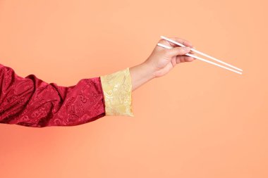 Yeni Çin yılın kutlu olsun. Kırmızı geleneksel cheongsam qipao ya da changshan elbisesi giyen Asyalı son sınıf öğrencisinin eli turuncu arka planda izole edilmiş beyaz çubuklarla el ele tutuşuyor..