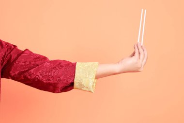 Yeni Çin yılın kutlu olsun. Kırmızı geleneksel cheongsam qipao ya da changshan elbisesi giyen Asyalı son sınıf öğrencisinin eli turuncu arka planda izole edilmiş beyaz çubuklarla el ele tutuşuyor..