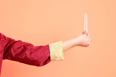 Yeni Çin yılın kutlu olsun. Kırmızı geleneksel cheongsam qipao ya da changshan elbisesi giyen Asyalı son sınıf öğrencisinin eli turuncu arka planda izole edilmiş beyaz çubuklarla el ele tutuşuyor..