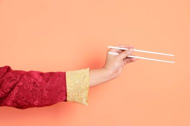 Yeni Çin yılın kutlu olsun. Kırmızı geleneksel cheongsam qipao ya da changshan elbisesi giyen Asyalı son sınıf öğrencisinin eli turuncu arka planda izole edilmiş beyaz çubuklarla el ele tutuşuyor..