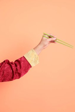 Yeni Çin yılın kutlu olsun. Kırmızı geleneksel cheongsam qipao ya da changshan elbisesi giyen Asyalı son sınıf öğrencisinin eli turuncu arka planda izole edilmiş yeşil çubuklarla el ele tutuşuyor..