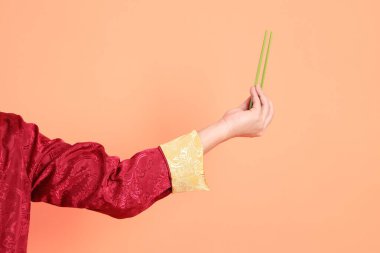 Yeni Çin yılın kutlu olsun. Kırmızı geleneksel cheongsam qipao ya da changshan elbisesi giyen Asyalı son sınıf öğrencisinin eli turuncu arka planda izole edilmiş yeşil çubuklarla el ele tutuşuyor..