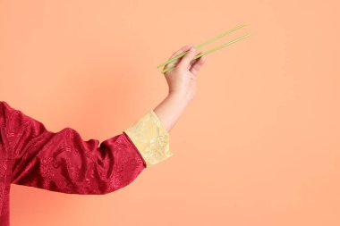 Yeni Çin yılın kutlu olsun. Kırmızı geleneksel cheongsam qipao ya da changshan elbisesi giyen Asyalı son sınıf öğrencisinin eli turuncu arka planda izole edilmiş yeşil çubuklarla el ele tutuşuyor..