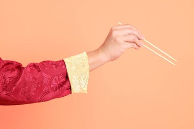 Yeni Çin yılın kutlu olsun. Kırmızı geleneksel cheongsam qipao ya da changshan elbisesi giyen Asyalı son sınıf öğrencisinin eli. Turuncu arka planda izole edilmiş ahşap çubukları tutan el hareketi..